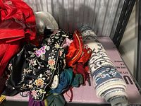 Vintage rain jackets in red, yellow, and blue, displayed with other fabrics and textiles including embroidered and floral patterns, and a rolled woven blanket or rug