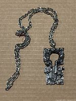 Full view of vintage Robert Larin pendant necklace with a rough textured abstract geometric pendant on silver chain lying on cardboard background.
