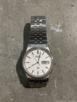 Top-down photo of a silver-tone Seiko wristwatch with stainless steel link band and a day-date display window. Watch is placed on a textured surface.