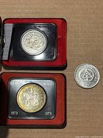 Three coins: one silver-colored coin in a protective case with an intricate floral pattern from 1876, one Canada dollar commemorative coin featuring RCMP on horseback from 1973 also in a protective case, and a loose silver coin commemorating the 1976 UN Habitat Conference with symbolic design.