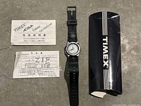 Timex Indiglo watch with black strap lying on the floor with instruction manuals and warranty card beside it, along with the original black Timex box.