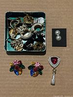 Overview photo of all vintage earrings in the lot placed on brown cardboard for scale and clarity, showing small blue and black box holding most earrings, colorful floral earrings, pearl studs and drop earring laid out beside the box.
