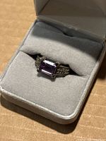 Ring displayed in a gray padded jewelry box showing geometric patterned band and rectangular purple gemstone set in prongs.
