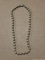 Full view of the vintage pearl necklace laid flat on a cardboard surface, showing the string of round pearls and the clasp at the top center.