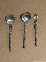 Three vintage sterling silver serving utensils displayed against cardboard: two spoons and one three-pronged fork, all with intricately embossed handles.