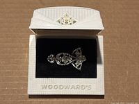 Silver tone metal brooch with clear rhinestones inside a white textured Woodwards branded box with black fabric interior.