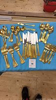 Complete set laid out on a blue cloth, showing all gold-toned cutlery pieces including knives, forks, spoons, and a serving fork.