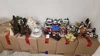 Wide view of assorted Christmas decorations arranged on cardboard boxes including ceramic and wooden Christmas trees, mini village houses, musical figurines, and Santa figurines.