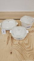 Three Corning Ware dishes with lids placed on wood floor, showing Blue Cornflower design on white ceramic body