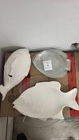 Three fish-shaped serving dishes on cardboard: a large white ceramic platter, a white ceramic soup tureen with fish shape, and a clear glass fish plate.