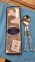 Raquel Breane 3-piece cake set in original box with clear acrylic handles and stainless steel blades, shown with a pair of salad servers with clear acrylic handles on blue fabric background