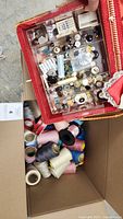 Photo showing an open box filled with various thread spools in assorted colors and a plastic organizer with buttons and sewing needles inside a red case.