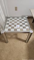 Side view of the vintage chessboard side table showing the metal frame and cylindrical legs on carpet.