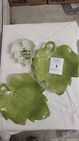 Three leaf-shaped dishes photographed on white cloth: two large green dishes with detailed leaf veining and one smaller white dish with floral design.