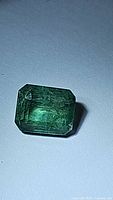 Photo of a deep green octagon cut natural emerald gemstone under direct light, showing luster and inclusions.