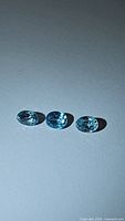 Three oval cut blue zircon gemstones arranged in a row on a neutral surface under light, showing clarity and color.