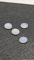 Four oval cabochon cut chalcedony stones laid on a textured black background showing their translucent, pearl white to lavender colors under diffuse light.