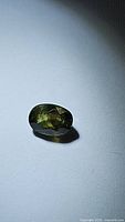 Photo of natural green sapphire gemstone, oval cut, approximately 6.8x5mm, showing facets and color under light on white background.