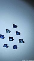 Eight princess cut blue sapphire gemstones laid out on a white surface showing their size and color variations.