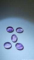 Photo showing five natural purple oval cabochon amethyst gemstones on a light background with visible internal inclusions.