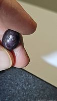 Close-up photo of an oval cabochon garnet held between fingers showing the gemstone's deep reddish-purple color and star effect reflection under light.