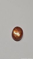 Natural oval sunstone cabochon with bright metallic flash, orange-copper color, on plain white background.