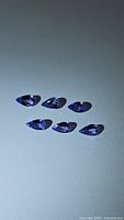 Six pear cut tanzanite gemstones arranged on a flat surface under light, showing their purple-blue color and clarity.