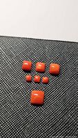 Seven polished cushion-cut red coral stones arranged on a textured black surface, showing size variation.