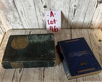 Lot of two antique books, with one blue embossed cover and one dark green cover with gold emblem, set against wooden backdrop.
