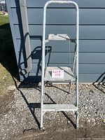 Front view of aluminum 2-step ladder leaning against gray siding, showing wide steps and rounded top handle.