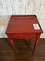 Closed view of the antique red wooden desk with a flat top panel and turned legs against a wooden wall.