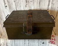 Full view of closed green metal locker box with leather strap securing the lid and black metal handles on the sides.