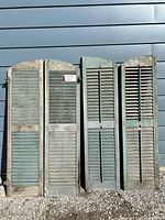 Front view of all four antique wooden shutters against wall showing two rectangular and two arched top shutters in aged blue-green paint with working slats and original hardware attached.