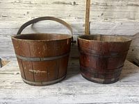 Two antique wooden buckets side by side on a wooden surface with a ruler in background for size reference.