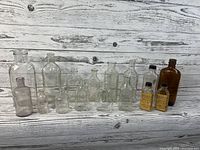 Wide view of all sixteen glass bottles arranged on a wooden surface, 15 clear glass and 1 amber bottle, including 2 bottles with labels and black caps.