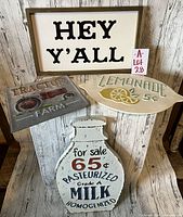 Four signs displayed against a wooden wall: wooden framed white 'Hey Y'ALL' sign, metal 'Lemonade 5¢' sign, metal milk bottle-shaped sign, and metal tractor sign.