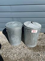 Two galvanized steel garbage cans with lids, placed side by side outdoors on gravel against a grey wall. Both show light weathering.