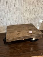 Full view of the handmade industrial cart coffee table with wooden plank top and cast iron wheels, placed on a wooden floor against a light patterned wall.