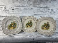 Three square porcelain plates with rounded corners laid flat, showing colorful cottage scenes at center with black and white floral borders and gold accents.