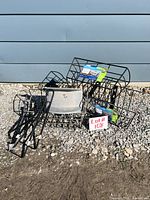 Photo showing the assortment of black metal hose hanger, wire baskets, and hanging brackets lying on gravel against a blue wall.