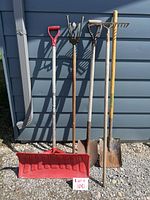 Photo of five outdoor garden tools against building wall: snow shovel (red plastic blade), cultivator, flat rake, round nose shovel, square nose shovel, all with wooden handles in used condition.