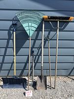 Photo shows six outdoor tools lined up against a grey wall on gravel ground. Tools include a snow shovel, fan rake, flat broom, square nosed shovel, edging shovel, and three-prong cultivator, all with wooden handles and varying metal or plastic working ends.