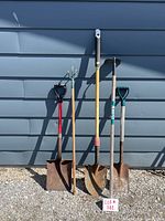 Photo of five outdoor garden tools lined up against a blue siding wall, showing varying shovel types, a cultivator, and a hoe.