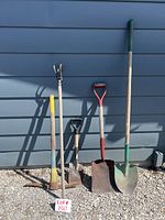Photo showing all five garden tools standing against a blue wall: pick axe, cultivator, small square nosed shovel, round nosed shovel, and long square nosed shovel.