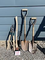 Five outdoor garden tools leaning against siding on gravel ground, including pruning shears, axe, two shovels, and hand cultivator, all with wooden handles and metal parts showing wear and rust.