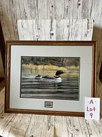 Framed print showing a loon on water, wood frame, white matting, with a certificate label below image.