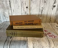 Three vintage cookbooks stacked on a wooden table with weathered white wood background. Titles visible on spines and front covers.