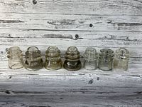 Seven vintage glass insulators arranged in a row on wood background, varied shapes and slight color differences.