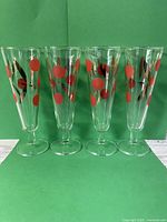 Set of four tall clear vintage glasses arranged side by side on a flat surface against a green background, showing red dot cherry and gold leaf pattern all around the exterior.