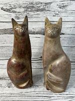 Front view of two hand-carved soapstone cat figurines, one reddish-brown and one light beige, placed side by side on a whitewashed wooden background.